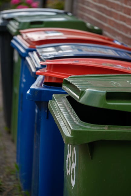 A close-up view of a tall, blue plastic rubbish bin with a partially open lid, positioned on a grassy outdoor area. The bin features a small, colorful sticker that reads 'FIGHT BACK!' in bold letters.